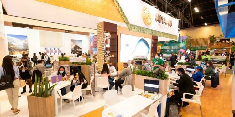 Stand, dorado y blanco, con mesas y sillas blancas en donde están sentadas las personas.