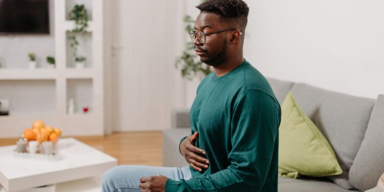In this image, a young man is seen sitting on a sofa in his living room, looking uncomfortable and holding his stomach, presumably due to digestive problems.