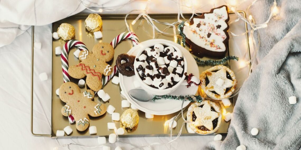 The the best (and worst) festive foods for sleep image shows a golden tray of gingerbread cookies, mince pies and marshmallow-topped hot chocolates placed on a bed dressed with faux fur blankets