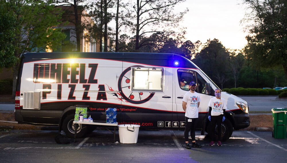Masked employees from the NHRMC Barto Center for Employee Fitness stand ready to deliver pizza slices from Wheelz Pizza to health care workers passing through the parking lot. (Port City Daily photo/Mark Darrough)