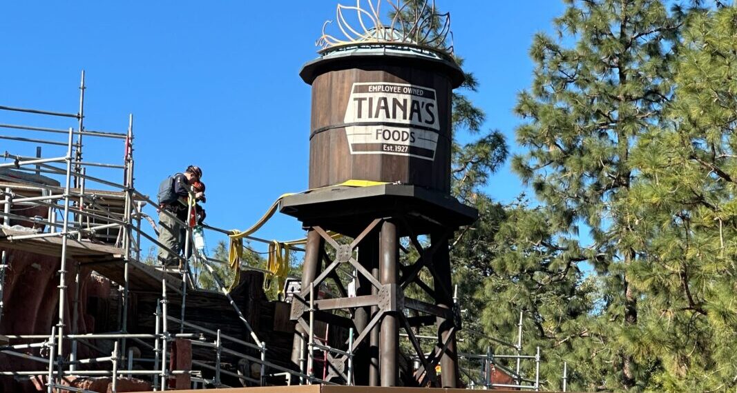 Two crew members work to the left of the Tiana's Foods water tower.