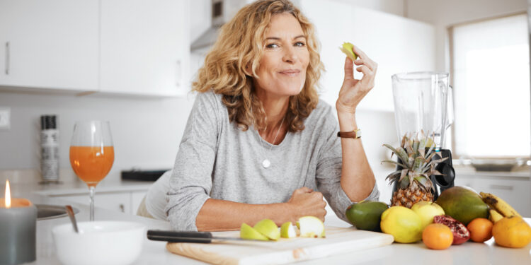 A woman in a grey top leaning over a counter with fresh fruit and a cutting board, eating cortisol-lowering foods