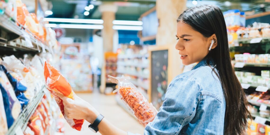 Dissatisfied young woman wearing casual outfit and earphones holding two packs of pasta looking at label while standing in grocery department in supermarket