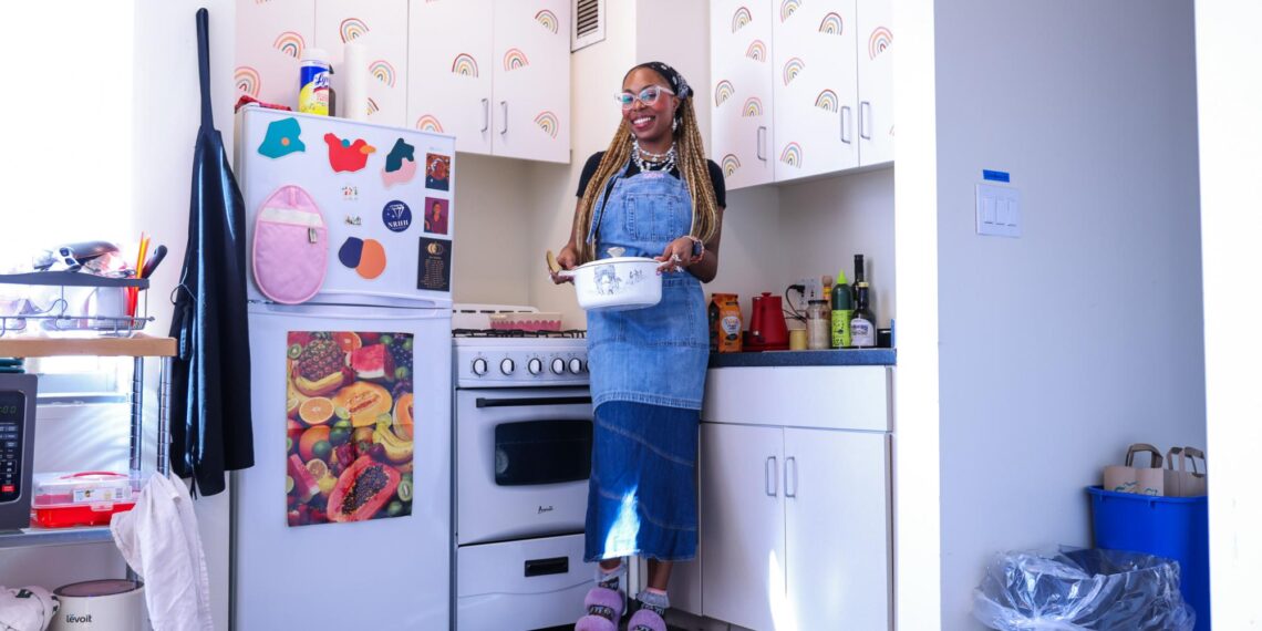 Sasha DuBose, in a blue apron and purple slippers, standing by a refrigerator and holding a white pot.
