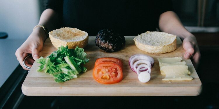 woman holding up wooden tray with lettuce, tomato, onions rings and cooked patty for burger