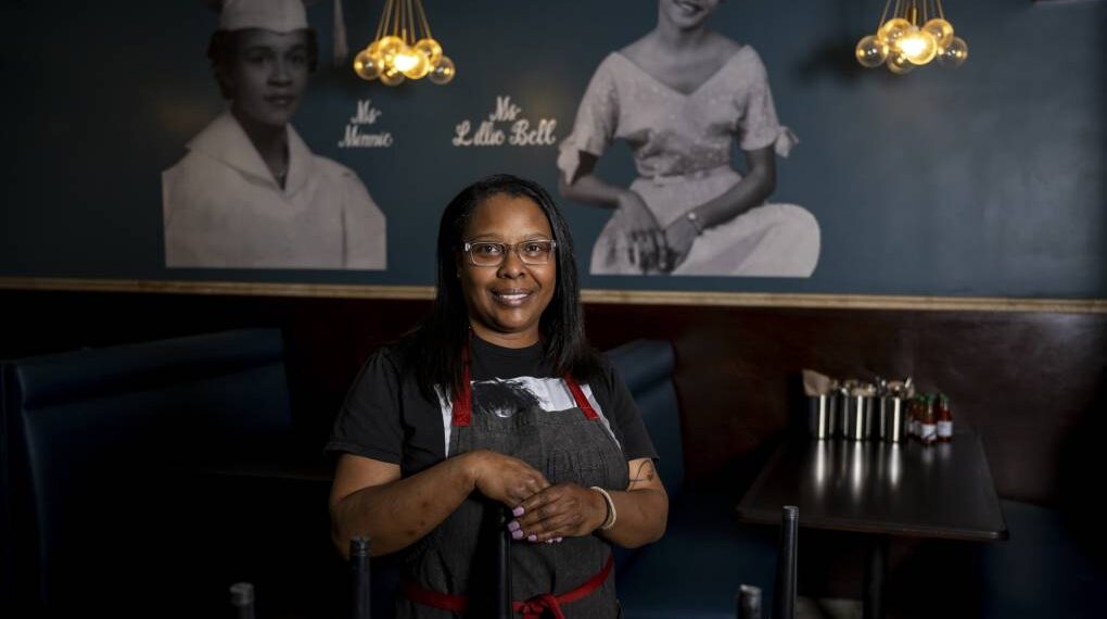 A chef poses inside her restaurant in front of a photo mural that shows her great-aunt and grandmother.