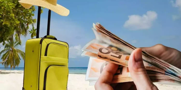Montaje con una imagen de fondo de una playa con una maleta y un sombrero de paja encima y en una esquina una mano contando billetes de 50 euros