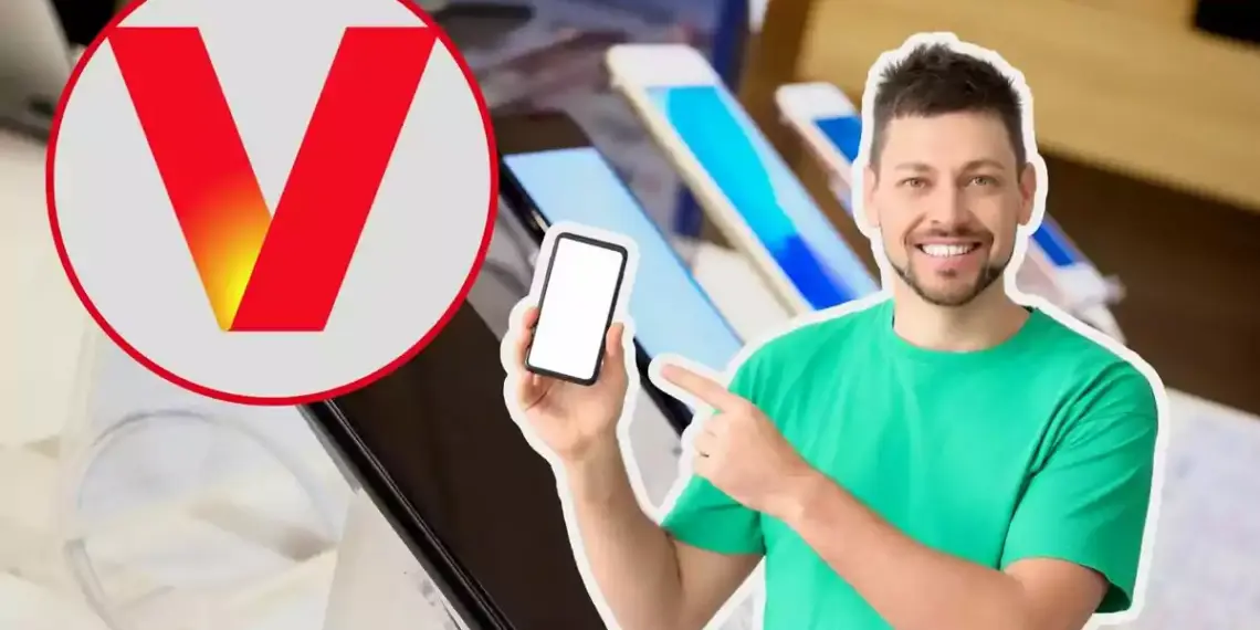 Un hombre sonriente con camiseta verde sostiene y señala un teléfono móvil con pantalla en blanco, junto a un logotipo circular con una letra "V" roja, en un entorno de tienda de tecnología con varios dispositivos en exhibición.