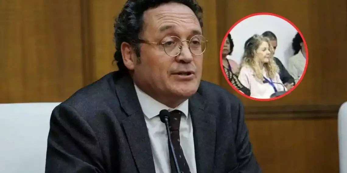 Un hombre con gafas y traje oscuro habla en un micrófono en una sala de conferencias con un recuadro que muestra a una mujer entre el público.