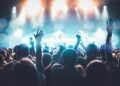 A lively crowd enjoys a concert, hands raised in excitement under bright stage lights.