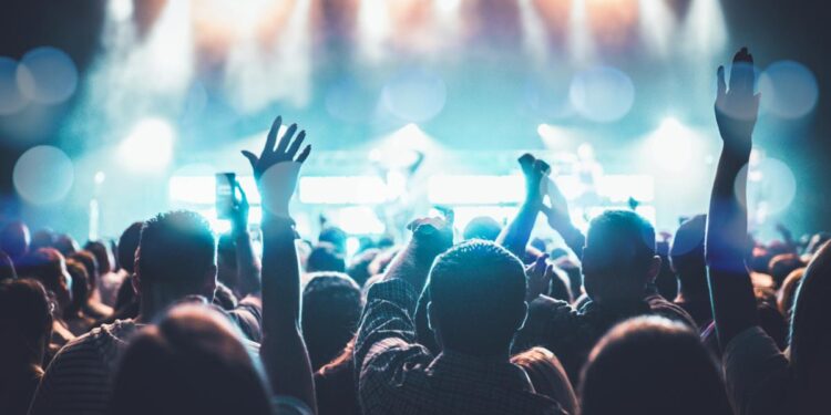 A lively crowd enjoys a concert, hands raised in excitement under bright stage lights.